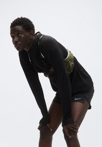 Atleta com camisa preta de manga comprida e calções pretos, usando um arnês atlético verde. O fundo é branco liso, destacando o equipamento.