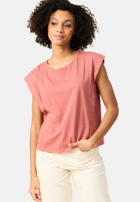 Vaaleanpunainen puuvillapaita, jossa on lyhyet, käännetyt hihat, pyöreä kaula-aukko ja rento malli, yhdistettynä kermaisiin housuihin.