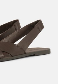 Brūna ādas plakanā sandale ar platajām krusteniskajām siksnām un aizmugures siksnu uz gludas pelēkas zolītes baltā fonā.