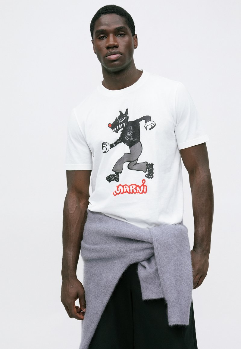 Witte T-shirt met een zwart cartoonfiguur met een rode neus en het woord "MARNI" in het rood. Model draagt een grijze trui om de taille geknoopt.