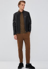 Zwarte leren jas met een staande kraag, bruine polo-shirt en bruine slim-fit broek. Zwarte suede schoenen maken de outfit compleet.