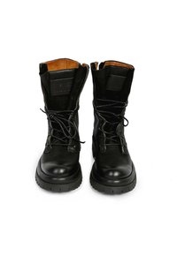 Paire de bottes de combat en cuir noir avec des lacets noués lâchement, semelles épaisses et doublure intérieure beige, posées côte à côte sur fond blanc.