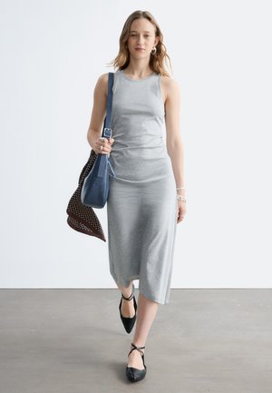 Młoda kobieta chodząca w pomieszczeniu, ubrana w szarą, bezrękawnikową sukienkę midi, czarne baleriny z wiązaniem przy kostce, srebrną biżuterię, niosąca niebieską torebkę ze brązową apaszką.