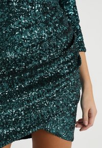 Tyrkysové třpytivé šaty s volnou tkaninou, vyznačující se vysokým výstřihem a dlouhými rukávy. Textura je lesklá a odrazivá s asymetrickým lemem.