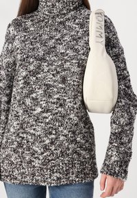 Gestrickter Pullover in Dunkelbraun und cremefarbenem Muster mit hohem Kragen. Hält eine weiße Tasche mit geprägtem "JEANS"-Tragegurt-Detail.