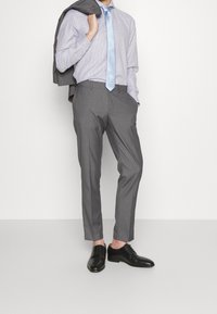Pantalon de costume gris et cravate bleu clair portés avec une chemise à carreaux. Des chaussures habillées noires complètent la tenue, avec un blazer gris posé sur une épaule.
