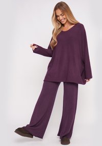 Übergroßer lila Pullover mit weiten Ärmeln, kombiniert mit passenden lila weit geschnittenen Hosen; aus weichem, strukturiertem Stoff. Braune Schuhe runden das Outfit ab.