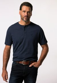 Maglietta henley a maniche corte blu navy con tre bottoni. Tessuto morbido, con un piccolo logo sul petto. Abbinata a jeans scuri e una cintura marrone.