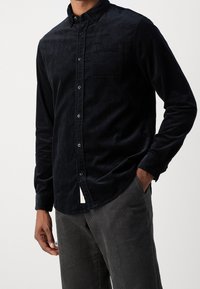 Camicia blu scuro in velluto a coste con bottoni e tasca, dotata di colletto classico e maniche lunghe, abbinata a pantaloni grigi in velluto a coste dalla texture distintiva.