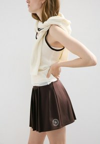 Mujer con top de punto sin mangas de color crema y falda plisada oscura con un logo circular bordado en el bajo, posando de lado con la mano en la cadera.