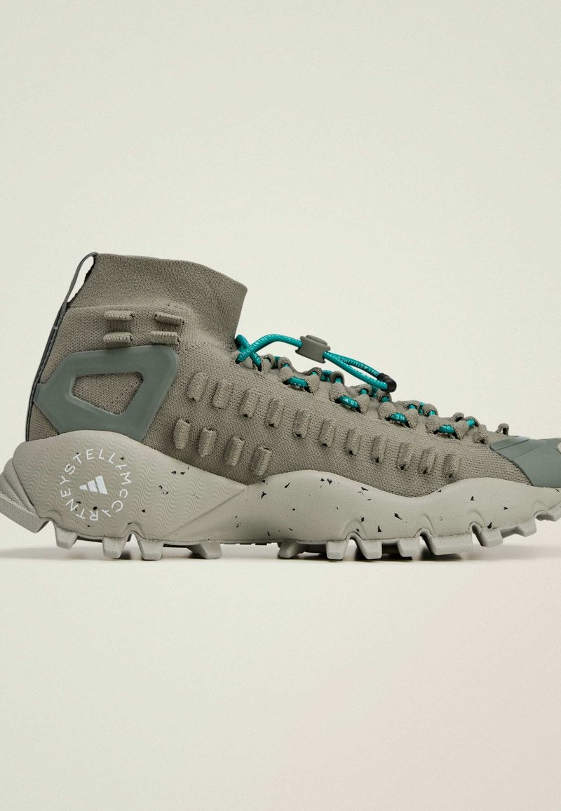 Lichtgrijze sportschoenen met een hoge schacht, gestructureerde bovenkant, turquoise veters en een robuuste zool met zwarte spikkels en profielnoppen.