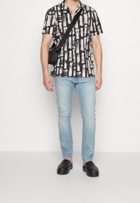 Kortärmad skjorta med ett mönster av svarta och krämfärgade penseldrag, kombinerad med ljusblå jeans och svarta slip-on skor.