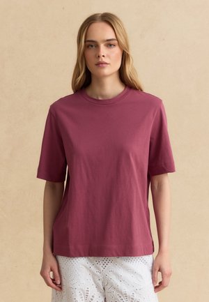 Vrouw met lang blond haar, die een effen mauve short-sleeve T-shirt en een witte eyelet kanten rok draagt, staande tegen een beige achtergrond.