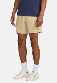 Pantalones cortos beige con aberturas laterales, tejido suave y un logotipo negro en el dobladillo izquierdo. Combinados con una camiseta azul marino y zapatillas blancas.
