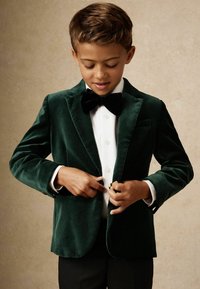 Blazer en velours vert avec finition satinée, présentant une fermeture à un bouton, un nœud papillon noir et une chemise blanche en dessous. Pantalons noirs visibles.