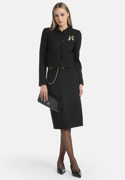 Black tailored jacket with button closure and pockets, paired with a pencil skirt. Accessories include a gold chain and a black crocodile-pattern clutch.