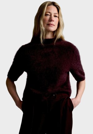 Donna con capelli biondi che indossa un maglione scuro e soffice a maniche corte e pantaloni scuri, in piedi con le mani nelle tasche di fronte a uno sfondo uniforme.