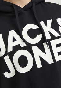 Schwarzer Kapuzenpullover mit großem weißen Text "JACK & JONES." Der Stoff ist weich und hat eine glatte Textur; er verfügt über verstellbare Kordeln.