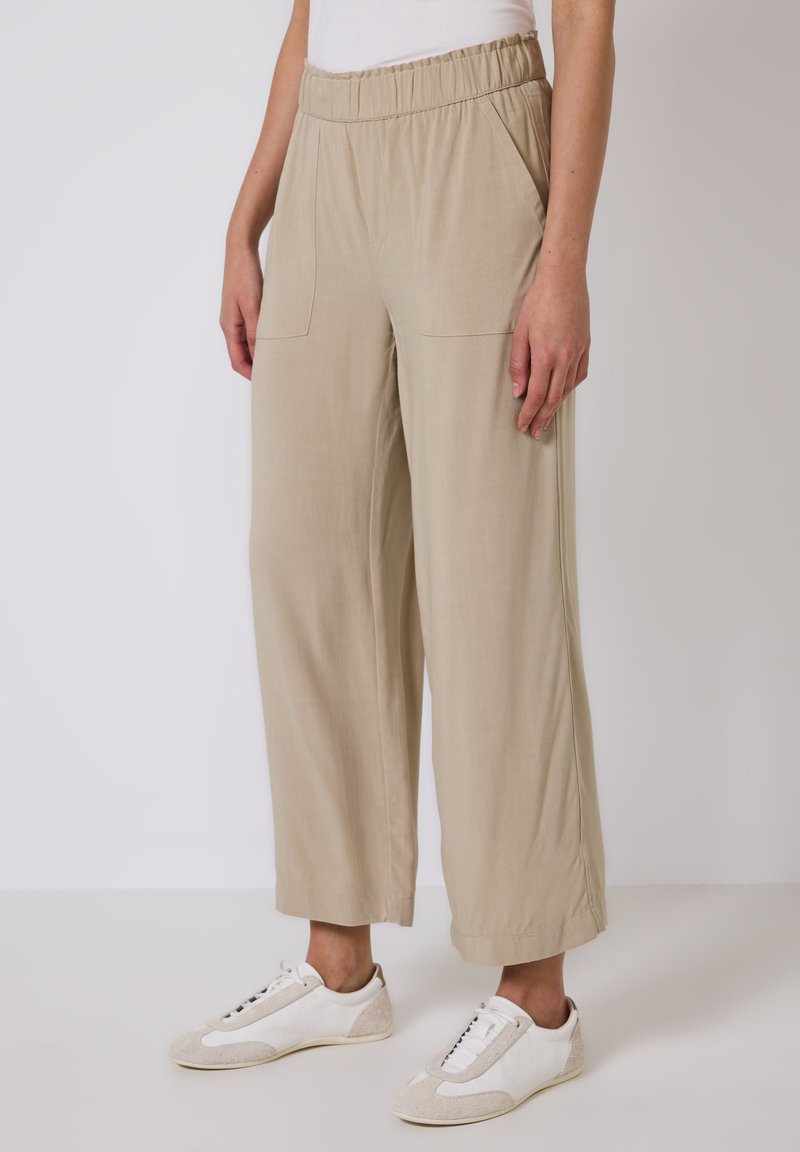 Beige weite Beinhosen mit elastischem Bund und zwei vorderen Taschen, kombiniert mit weißen und beigen Sneakern. Glatte Stoffstruktur.