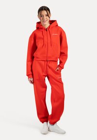 Rode cropped hoodie met rits en voorzakken, gecombineerd met bijpassende sweatpants. Beide hebben een gladde, zachte textuur en elastische manchetten.