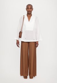 Blusa bianca a maniche lunghe con scollatura a V e cordoncino. Abbinata a pantaloni marroni a gamba larga. La modella tiene una piccola borsa a tracolla marrone.