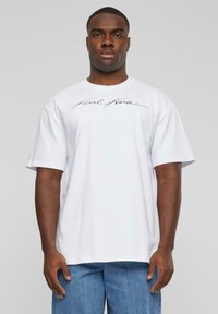 T-shirt en coton blanc avec un col rond et des manches courtes, arborant un logo noir écrit à la main sur la poitrine. Ourlet droit, coupe décontractée.
