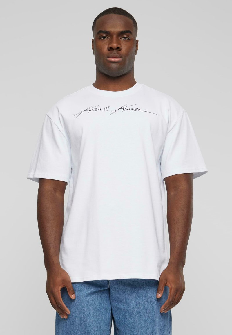 T-shirt en coton blanc avec un col rond et des manches courtes, arborant un logo noir écrit à la main sur la poitrine. Ourlet droit, coupe décontractée.