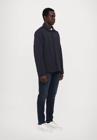 Marineblau gesteppte Jacke mit Knopfverschluss, klassischem Kragen, kombiniert mit dunklen Jeans und weißen Sneakers, vor einem schlichten Hintergrund stehend.