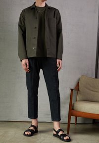 Giacca verde scuro con taschino sul petto, abbinata a una maglietta coordinata e pantaloni in denim scuri, completata da sandali neri con cinturini.