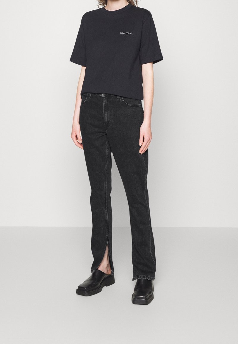 Black cotton t-shirt with short sleeves, paired with high-waisted black jeans featuring side slits at the ankles, and black footwear.