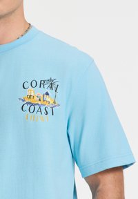 Svijetloplava pamučna majica s višebojnim grafičkim dizajnom plažnih koliba, palmi i natpisom 'Coral Coast' u crnoj i žutoj boji.