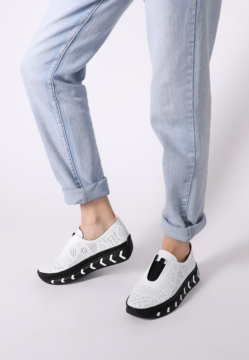 Weiße Slipper aus perforiertem Material, schwarze dicke Sohle mit weißen Chevron-Akzenten und elastischen Seiteneinsätzen für zusätzlichen Komfort.
