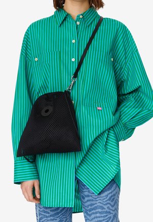 Personne portant une chemise oversize à rayures vertes et bleues, un pantalon bleu à motifs et un sac bandoulière en maille noire.