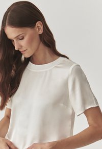 Bílá blůzka s krátkým rukávem vyrobená z hladkého materiálu, s kulatým výstřihem a jemným leskem, se minimalistickým designem a bez viditelných vzorů.