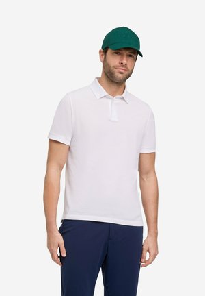 Mann trägt ein weißes kurzärmeliges Poloshirt, marineblaue Hose und eine dunkelgrüne Baseballkappe und steht vor weißem Hintergrund.