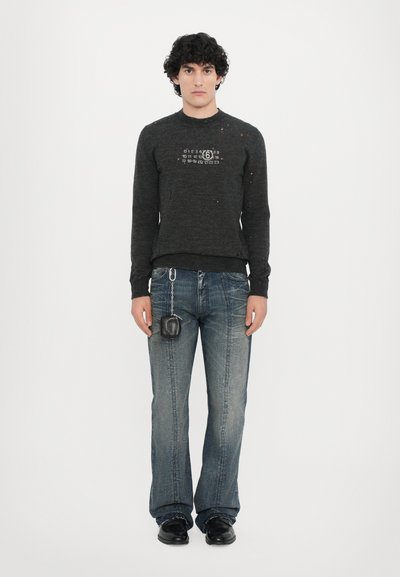 Dunkelgrauer Pullover mit Textdesign und Farbspritzern. Blaue schmal geschnittene Jeans mit dezenten Verfärbungen. Kleine schwarze Tasche am Gürtelschlaufen befestigt. Schwarze Schuhe.