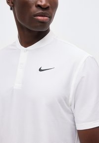 Κοντινό πλάνο ενός ατόμου που φοράει λευκό κοντομάνικο πουκάμισο Nike με γιακά δύο κουμπιών και μαύρο λογότυπο Nike στο στήθος.