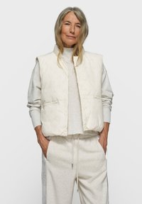 Gepolsterte, ärmellose Weste in Off-White mit dezentem Ton-in-Ton-Muster. Kombiniert mit hellgrauen, bequemen Jogginghosen mit elastischem Bund.