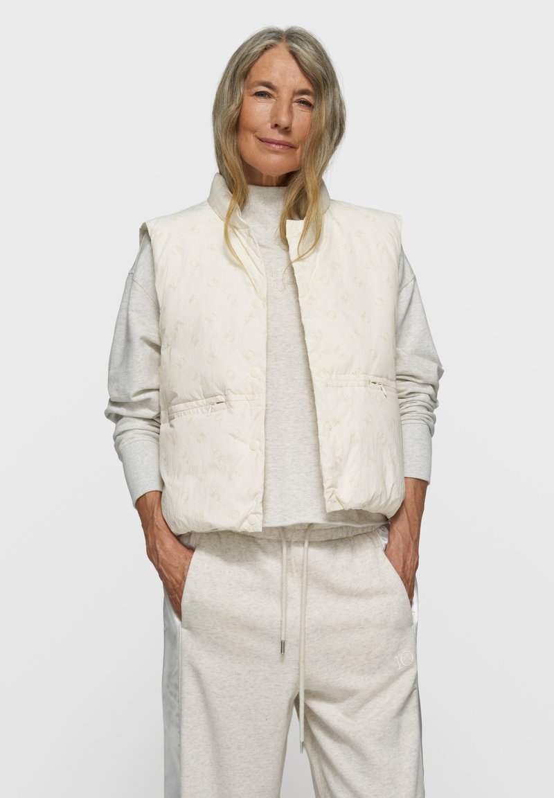 Gepolsterte, ärmellose Weste in Off-White mit dezentem Ton-in-Ton-Muster. Kombiniert mit hellgrauen, bequemen Jogginghosen mit elastischem Bund.