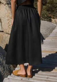 Jupe maxi en lin noir avec une taille élastique, tombant jusqu'aux chevilles. Portée avec des sabots à enfiler de couleur marron clair ornés de clous.