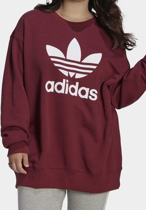 Personne portant un sweat Adidas bordeaux ample avec un grand logo blanc et un pantalon gris, partiellement visible, sur un fond uni.