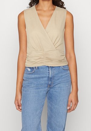 Woman wearing sleeveless beige ruched wrap top and light blue high-waisted jeans, standing against a plain background.