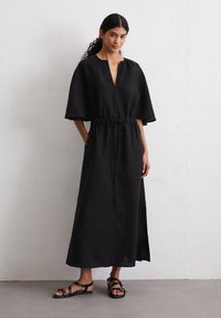 Abito maxi in lino nero con scollo a V, maniche corte e larghe, e vita con cordoncino. Il tessuto presenta una sottile texture e spacchi laterali. Indossato con sandali piatti.