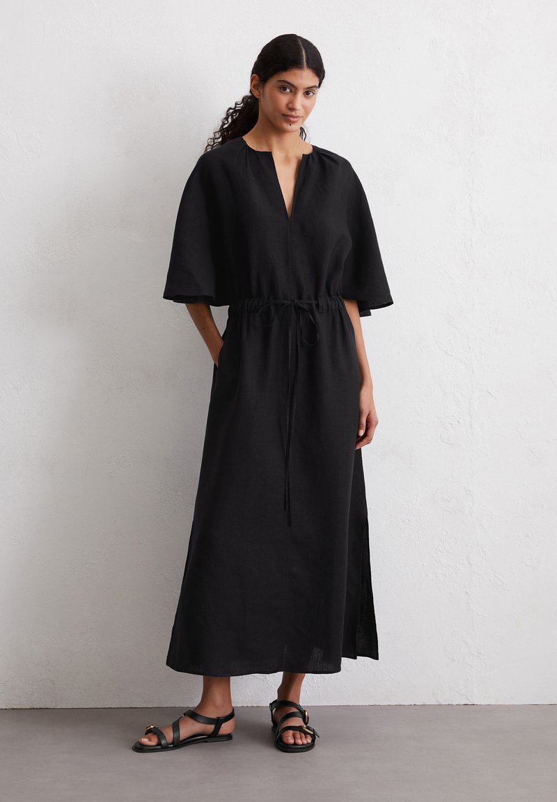 Abito maxi in lino nero con scollo a V, maniche corte e larghe, e vita con cordoncino. Il tessuto presenta una sottile texture e spacchi laterali. Indossato con sandali piatti.