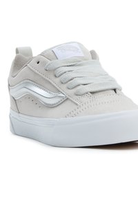 Jasnoszare zamszowe sneakersy z białą gumową podeszwą, z metaliczną srebrną paskiem po bokach i płaskimi szarymi sznurowadłami. Logo na języku.