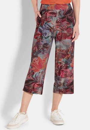 Vrouw in een cropped wijde broek met meerkleurige bloem- en bladpatronen, gecombineerd met witte sneakers en een gestructureerde oranje top.