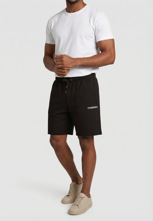 Man draagt een wit T-shirt met korte mouwen, zwarte shorts met trekkoord en het logo "CHABRAND", en beige sneakers, staand tegen een effen achtergrond.