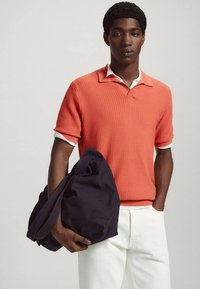 Jeune homme portant une chemise corail texturée à manches courtes par-dessus une chemise blanche à col, tenant un grand sac bleu foncé avec une main dans les poches.