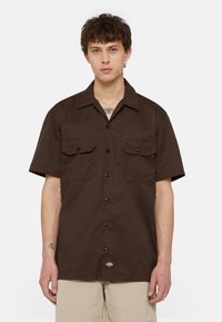 Brown short-sleeve shirt with two chest pockets, button front, and a patch logo at the hem. Made of lightweight fabric.