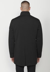 Cappotto nero con colletto alto, silhouette dritta, texture liscia e senza chiusure visibili. Design semplice senza motivi o decorazioni.